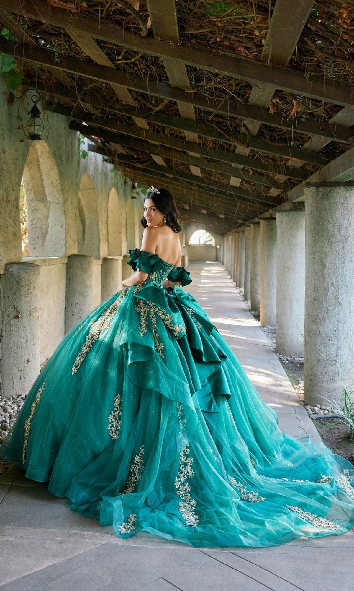 Wearing the Quinceanera Dress JT1504J by Juliet—a green ball gown with gold details, a corset bodice, and a glitter tulle skirt—a woman stands on a stone walkway beneath a wooden pergola, glancing back over her shoulder.
